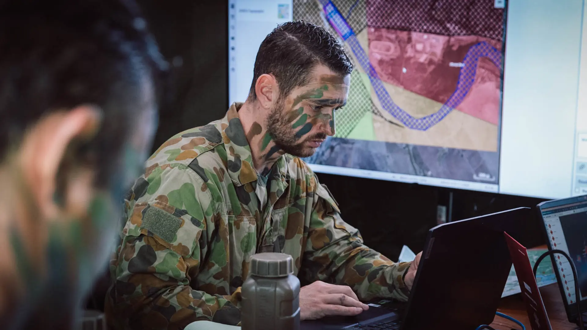 Camouflaged military staff working with mission data in SitaWare HQ software, analyzing a tactical map on laptops and a large screen inside a command post.